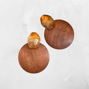 Puede incluir: Un par de pendientes con una base redonda de madera y una piedra translúcida de color ámbar. La madera tiene un rico tono marrón y la piedra un tono dorado. Los pendientes tienen un diseño sencillo pero elegante, con una paleta de colores naturales.