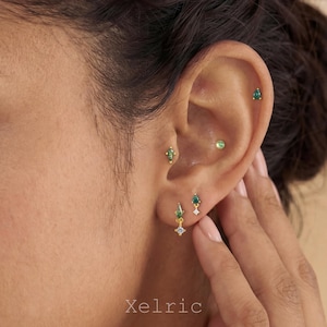 Puede incluir: Primer plano de una oreja adornada con múltiples pendientes dorados. Los pendientes presentan piedras preciosas verdes esmeralda y transparentes en varias formas, incluyendo lágrimas y tachuelas. La marca "Xelric" es visible en la parte inferior.