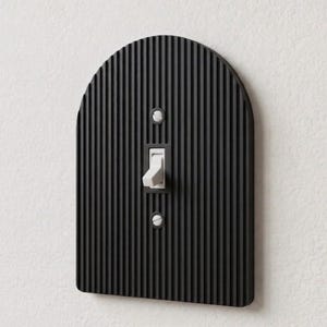 May include: A black arched light switch plate with a vertical ribbed texture. The plate features a central rectangular cutout for a white toggle switch and two circular openings for mounting screws, which are visible with white screw heads.