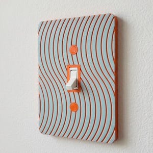 May include: A decorative light switch cover with a wavy pattern in light blue and orange. The switch itself is white, set in an orange frame. Two orange circles are at the top and bottom of the cover.