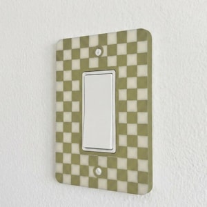 May include: A decorative light switch cover with a checkered pattern in olive green and cream. The cover has a rectangular shape with a single white rocker switch in the center. The cover is mounted on a white wall.
