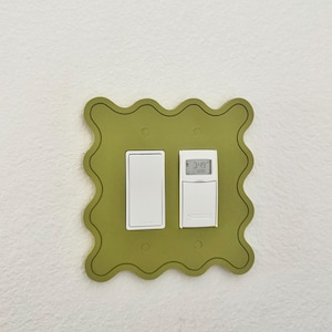 May include: Olive green light switch plate with a wavy edge, holding a white light switch and a digital thermostat. The thermostat displays the time. The plate is mounted on a white textured wall.