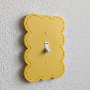 May include: A bright yellow light switch cover plate with a scalloped, cloud-like edge. The rectangular plate features a central vertical toggle switch in white and two small circular screw holes positioned above and below the switch mechanism.
