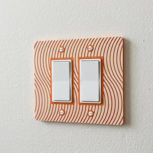 May include: A double light switch wall plate featuring a light peach background with a pattern of thin, wavy, dark orange lines. The rectangular plate is secured with four white screws and contains two white rocker-style light switches.