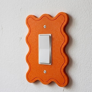 Peut inclure: Un cache-interrupteur orange aux bords ondulés avec un interrupteur à bascule blanc. Le cache est monté sur un mur blanc avec deux vis. L'interrupteur est en position marche.