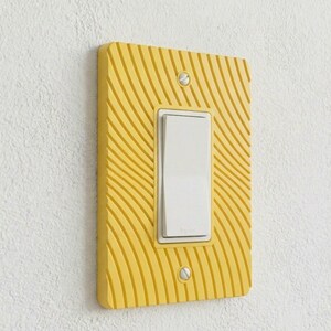 May include: A rectangular yellow light switch wall plate featuring a textured pattern of curved, radiating lines. The plate is secured with two silver screws and surrounds a white rocker-style light switch with the brand name Legrand printed on it.