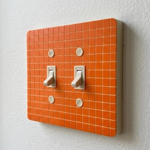 May include: A double light switch wall plate covered in a grid of small, bright orange square tiles. The plate features two off-white toggle switches positioned in the center, each flanked by two small, circular off-white screw covers.