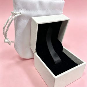 May include: A white jewelry box with a black interior and a black ribbon, next to a white drawstring pouch. The box is open, revealing the interior. The background is a solid pink color.
