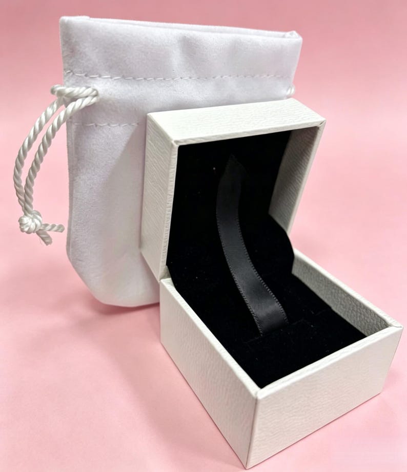 May include: A white jewelry box with a black interior and a black ribbon, next to a white drawstring pouch. The box is open, revealing the interior. The background is a solid pink color.