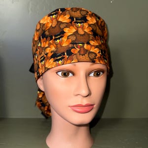 May include: A patterned scrub cap featuring a repeating design of cartoon turkeys in hats with orange and brown feathers. The cap is tied in the back with a black ribbon. The background is a muted green.