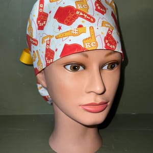 May include: White surgical scrub hat with a red and yellow print of pointing hands and the words "GO TEAM #1 KC".