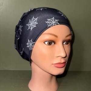 May include: A black scrub cap featuring a repeating white spiderweb pattern. The fabric is gathered at the back, providing a fitted look. The cap covers the crown of the head and is made of a smooth, lightweight material with a consistent print throughout.