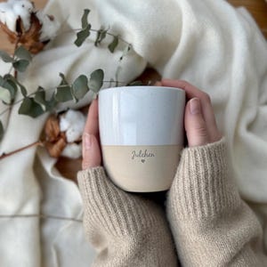 Könnte beinhalten: Eine zweifarbige Keramik-Tasse mit einem gesprenkelten weißen oberen Teil und einem beigefarbenen unteren Teil. Das Wort "Julchen" ist in einer Schreibschrift auf dem beigefarbenen Teil geschrieben, mit einem kleinen Herzsymbol darunter. Die Tasse wird von einer Person gehalten, die einen beigefarbenen Pullover trägt.