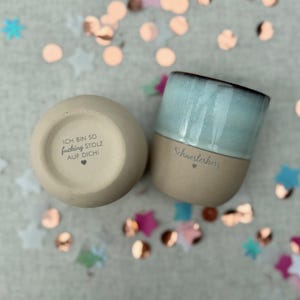 Könnte beinhalten: Zwei Keramikbecher mit Text. Ein Becher ist hellbeige mit dem Text "ICH BIN SO fucking STOLZ AUF DICH!" und einem Herz. Der andere Becher ist unten hellbeige und oben hellblau mit dem Text "Schwesterherz" und einem Herz.