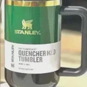 Puede incluir: Un vaso Stanley Quencher H2.0 verde y negro con un asa negra. El vaso tiene una banda dorada y el logotipo de Stanley. La etiqueta dice "QUENCHER H2.0 TUMBLER" y tiene una capacidad de 1,18 L.