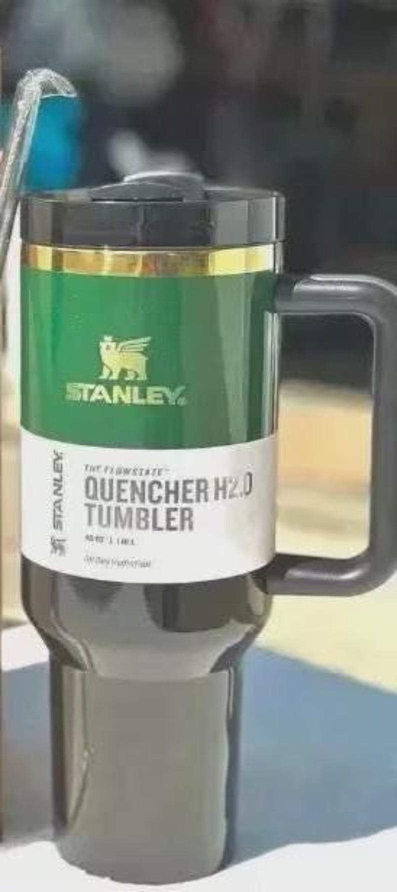 Puede incluir: Un vaso Stanley Quencher H2.0 verde y negro con un asa negra. El vaso tiene una banda dorada y el logotipo de Stanley. La etiqueta dice "QUENCHER H2.0 TUMBLER" y tiene una capacidad de 1,18 L.