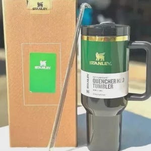 Puede incluir: Un vaso Stanley Quencher H2.0 verde y negro con asa, pajita y caja de cart&oacute;n. El vaso tiene una banda dorada y el logotipo de Stanley. La caja tiene el logotipo de Stanley y una inserci&oacute;n verde.