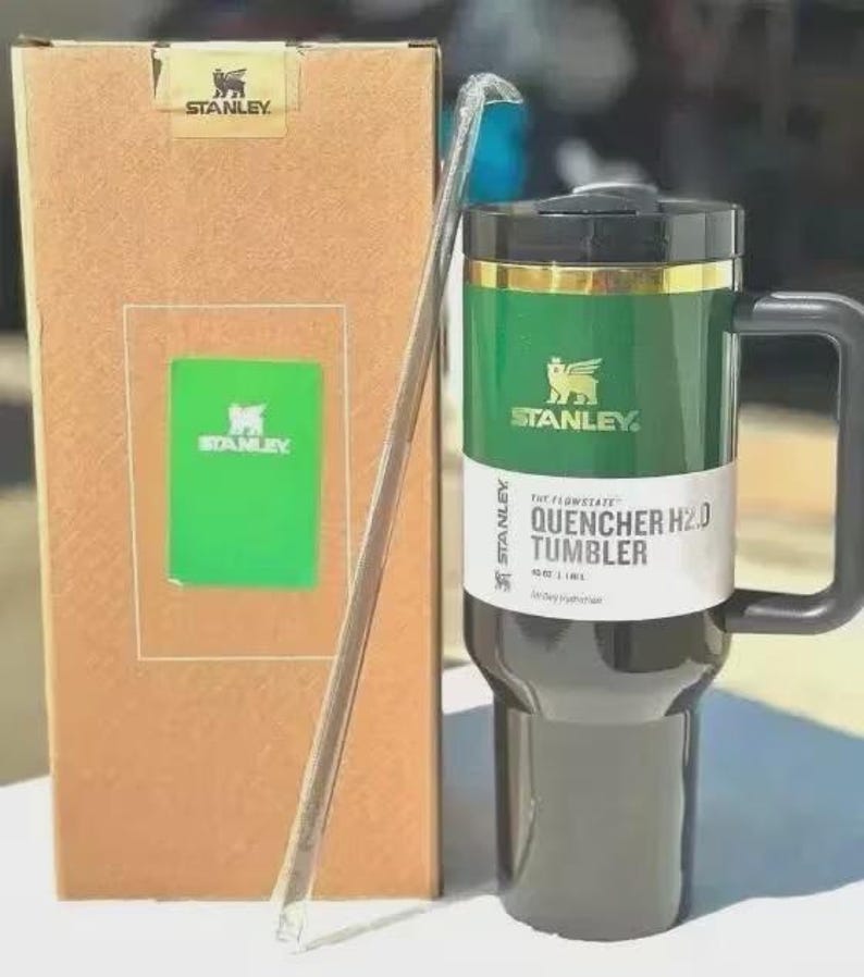 Puede incluir: Un vaso Stanley Quencher H2.0 verde y negro con asa, pajita y caja de cart&oacute;n. El vaso tiene una banda dorada y el logotipo de Stanley. La caja tiene el logotipo de Stanley y una inserci&oacute;n verde.