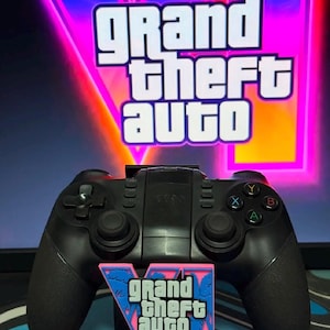 Op de afbeelding: Een zwarte draadloze videogamecontroller met twee analoge sticks, een richtingspad en vier gekleurde actieknoppen met de letters A, B, X en Y. Een klein roze en blauw Grand Theft Auto VI logo-embleem is aan de voorzijde van de controller bevestigd.