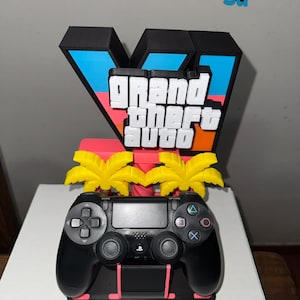 Puede incluir: Un mando de videojuego negro sobre un soporte con un gran letrero en forma de V con el texto Grand Theft Auto en letras blancas. El soporte incluye dos siluetas de palmeras amarillas y una barra horizontal rosa sobre un fondo negro.