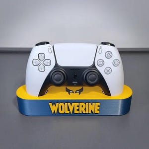 May include: A white and black video game controller resting on a yellow and blue display stand. The stand features the text WOLVERINE in yellow letters on the front, along with a black graphic icon of a mask positioned below the controller.