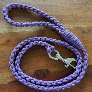 May include: A braided lavender dog lead with a silver metal clasp. The lead is made of a thick, durable material. The lead is looped and rests on a wooden surface. Designed for pet owners.