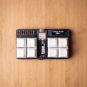 May include: A black rectangular macro keypad featuring eight white square keycaps, a small digital screen displaying the text SimPad Drums type..., a Seeed Studio controller board with a red LED, and a series of small electronic components.