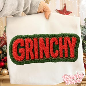 May include: A white fabric item with the word "GRINCHY" in large, red, textured letters. The letters are outlined with a green border and a gold-colored trim. The item is held up by a person.