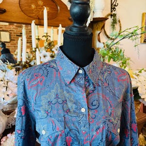 May include: A blue button-down shirt with a paisley pattern in red, gray, and gold. The shirt has a collared neckline, two chest pockets, and white buttons. The shirt is displayed on a black mannequin head.