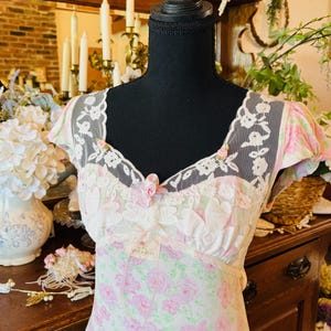 May include: A vintage-style, floral-print dress with a delicate lace neckline and cap sleeves. The dress features a soft pink and green rose pattern on a white background. A small pink bow accents the neckline. The dress is displayed on a black mannequin.
