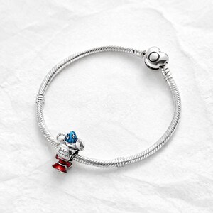 Puede incluir: Una pulsera de plata con un cierre en forma de coraz&oacute;n. La pulsera presenta un charm con un dise&ntilde;o rojo, blanco y azul, con las palabras "Believe in magic" inscritas. El charm tiene forma de trofeo.