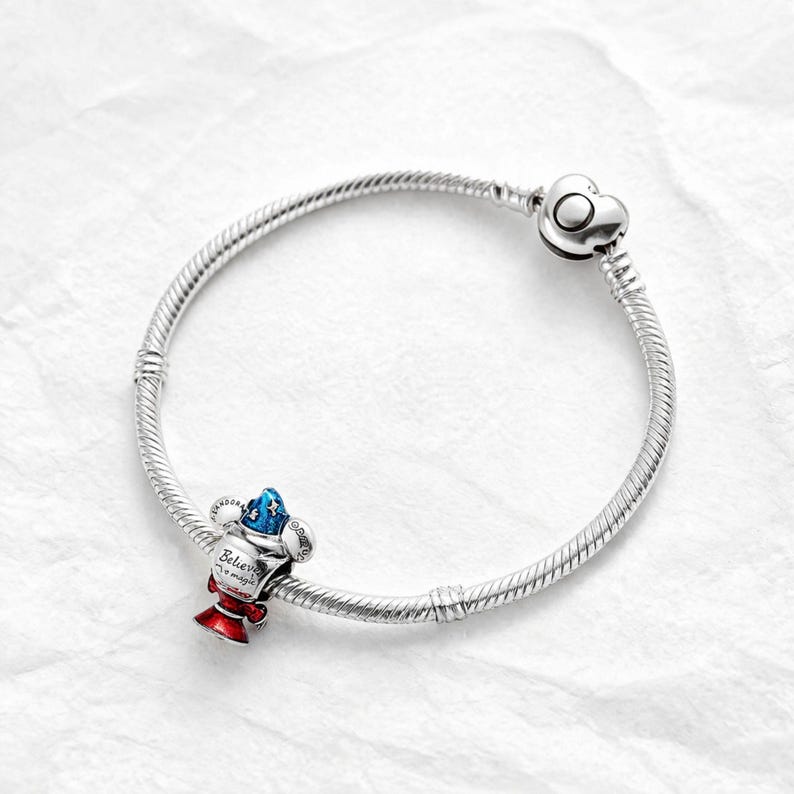 Puede incluir: Una pulsera de plata con un cierre en forma de coraz&oacute;n. La pulsera presenta un charm con un dise&ntilde;o rojo, blanco y azul, con las palabras "Believe in magic" inscritas. El charm tiene forma de trofeo.