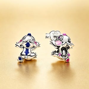 May include: Two silver charms featuring a cartoon character with blue, pink, and white accents. One charm has arms outstretched, the other holds a heart-shaped object. The charms are on a gold background.