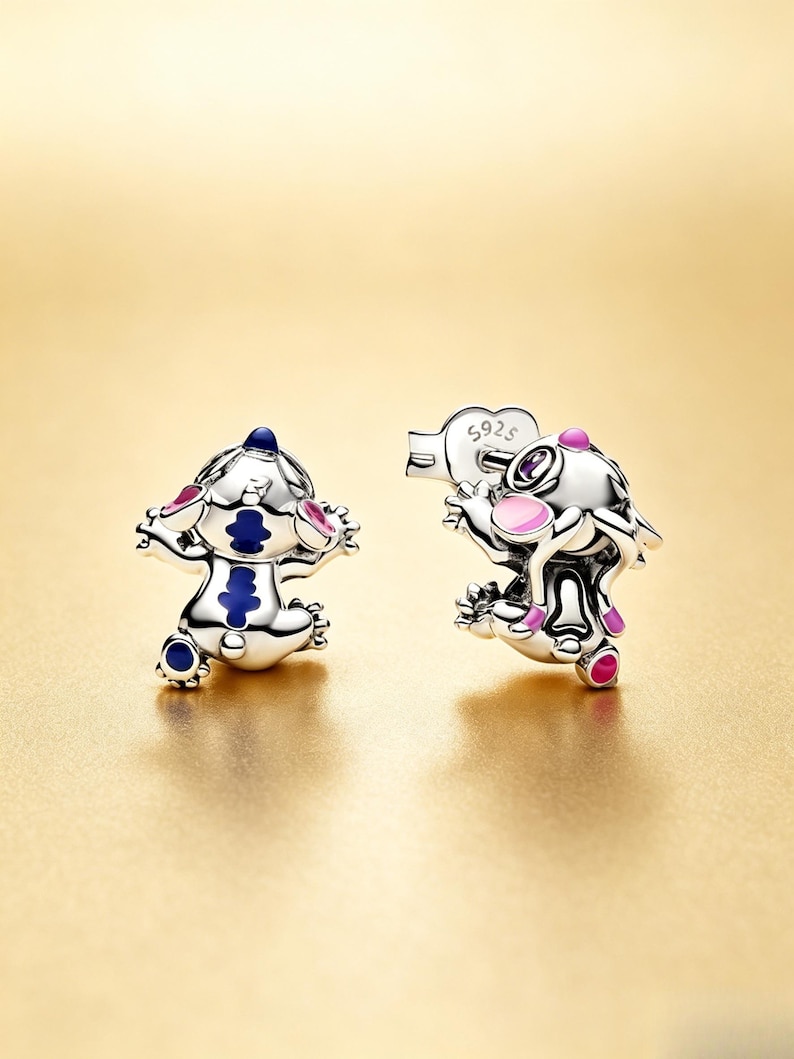 May include: Two silver charms featuring a cartoon character with blue, pink, and white accents. One charm has arms outstretched, the other holds a heart-shaped object. The charms are on a gold background.