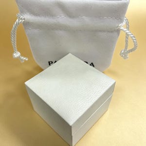 May include: A white, textured square box with a matching drawstring pouch. The box is closed and sits in front of the pouch. The pouch has a white drawstring closure. The box and pouch are on a light yellow surface.