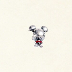 May include: A silver charm in the shape of a mouse with large ears, a red bow, and small feet. The charm has the text "Disney" and "925 ALE" engraved on it. The charm is a collectible item.