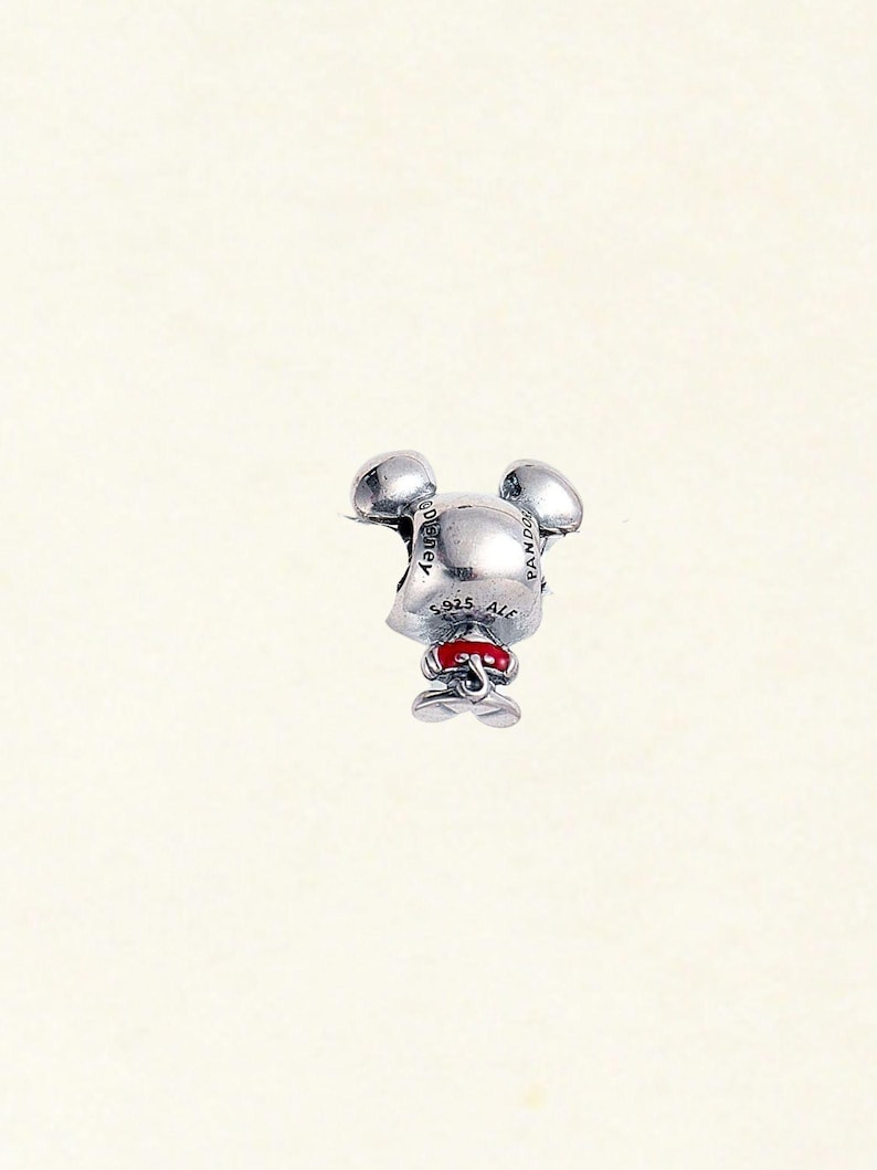 May include: A silver charm in the shape of a mouse with large ears, a red bow, and small feet. The charm has the text "Disney" and "925 ALE" engraved on it. The charm is a collectible item.