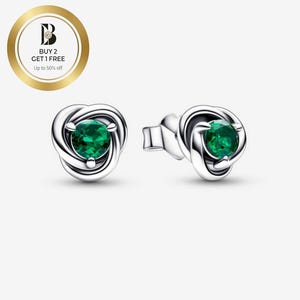 Puede incluir: Pendientes de plata con dise&ntilde;o de nudo, cada uno con una piedra preciosa verde redonda y vibrante. Los pendientes se muestran sobre un fondo claro. Un emblema circular dorado con texto est&aacute; en la esquina superior izquierda.