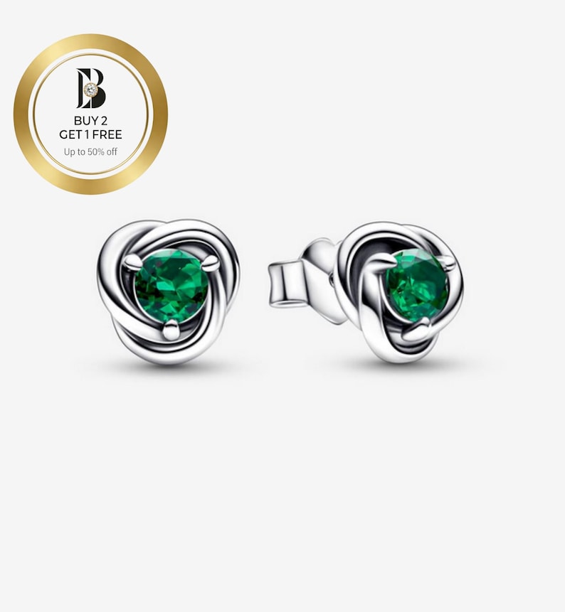 Puede incluir: Pendientes de plata con dise&ntilde;o de nudo, cada uno con una piedra preciosa verde redonda y vibrante. Los pendientes se muestran sobre un fondo claro. Un emblema circular dorado con texto est&aacute; en la esquina superior izquierda.