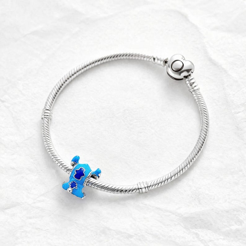 May include: A silver charm bracelet with a heart-shaped clasp. A blue and white enamel charm in the shape of a cartoon character is attached to the bracelet. The bracelet is on a white surface.