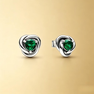 Puede incluir: Un par de pendientes de bot&oacute;n plateados. Cada pendiente presenta un dise&ntilde;o de nudo con una brillante gema verde esmeralda redonda en el centro. Los pendientes est&aacute;n sobre un fondo dorado suave.