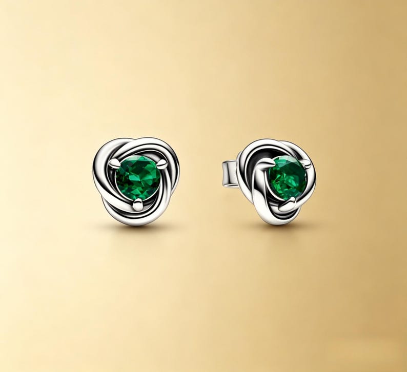 Puede incluir: Un par de pendientes de bot&oacute;n plateados. Cada pendiente presenta un dise&ntilde;o de nudo con una brillante gema verde esmeralda redonda en el centro. Los pendientes est&aacute;n sobre un fondo dorado suave.