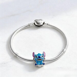 May include: A silver charm bracelet with a blue, pink, and white Stitch charm. The charm features Stitch, a character from the animated film Lilo & Stitch. The bracelet has a round clasp with the word "Pandora" on it.