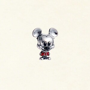 May include: A silver-colored charm depicting Mickey Mouse. The charm features Mickey's iconic large ears, a black nose, and a red and white polka-dotted outfit. The charm is a miniature representation of the beloved cartoon character.