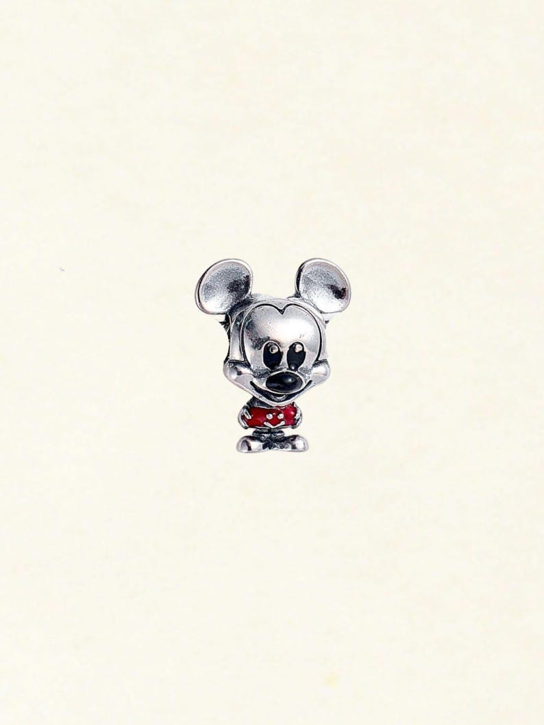 May include: A silver-colored charm depicting Mickey Mouse. The charm features Mickey's iconic large ears, a black nose, and a red and white polka-dotted outfit. The charm is a miniature representation of the beloved cartoon character.