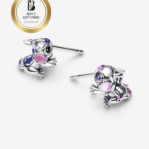 May include: A pair of silver-colored stud earrings featuring a cartoon character design. The earrings have blue and pink accents. The image also includes a gold-colored circle with text that reads "BUY 2 GET 1 FREE Up to 50% off."