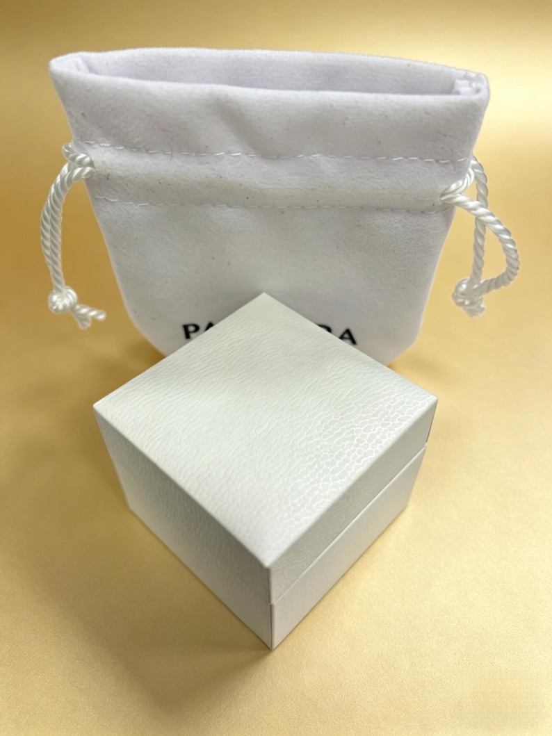 May include: A white, textured square box with a matching drawstring pouch. The box is closed and sits in front of the pouch. The pouch has a white drawstring closure. The box and pouch are on a light yellow surface.