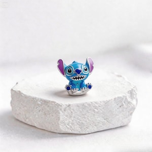 May include: A blue and silver charm featuring the character Stitch from the animated film Lilo & Stitch. The charm has a smiling expression with white teeth, large ears, and blue feet. The charm is set on a white, textured surface.