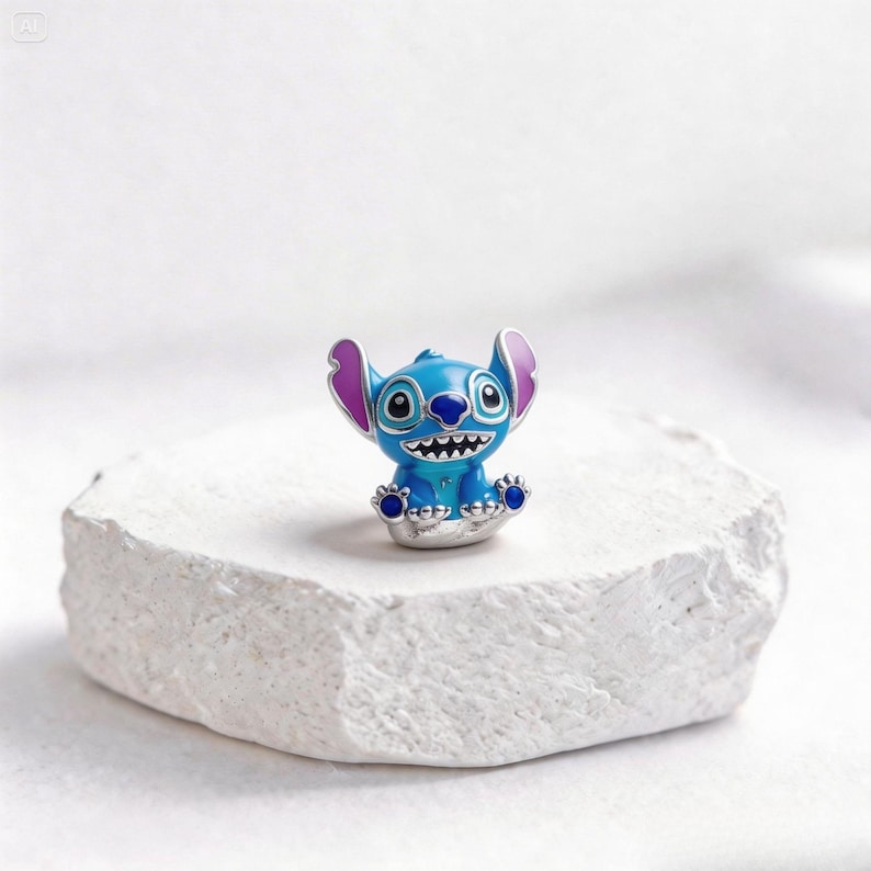 May include: A blue and silver charm featuring the character Stitch from the animated film Lilo & Stitch. The charm has a smiling expression with white teeth, large ears, and blue feet. The charm is set on a white, textured surface.