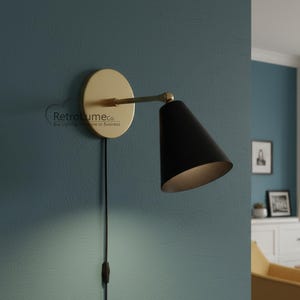 Op de afbeelding: Zwarte en gouden wandlamp met een conische kap. De lamp heeft een ronde, gouden basis en een zwart snoer met een schakelaar. De muur is blauwgroen. De lamp is van Retrolume co.